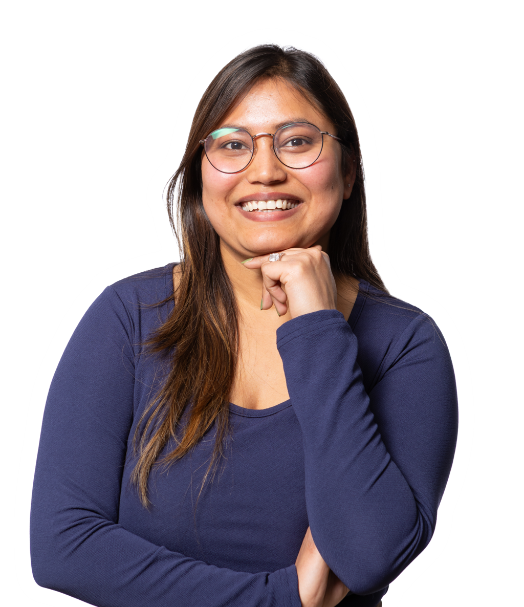 A woman with glasses smiles and rests her chin on her hand. She is wearing a blue shirt and brown pants.
