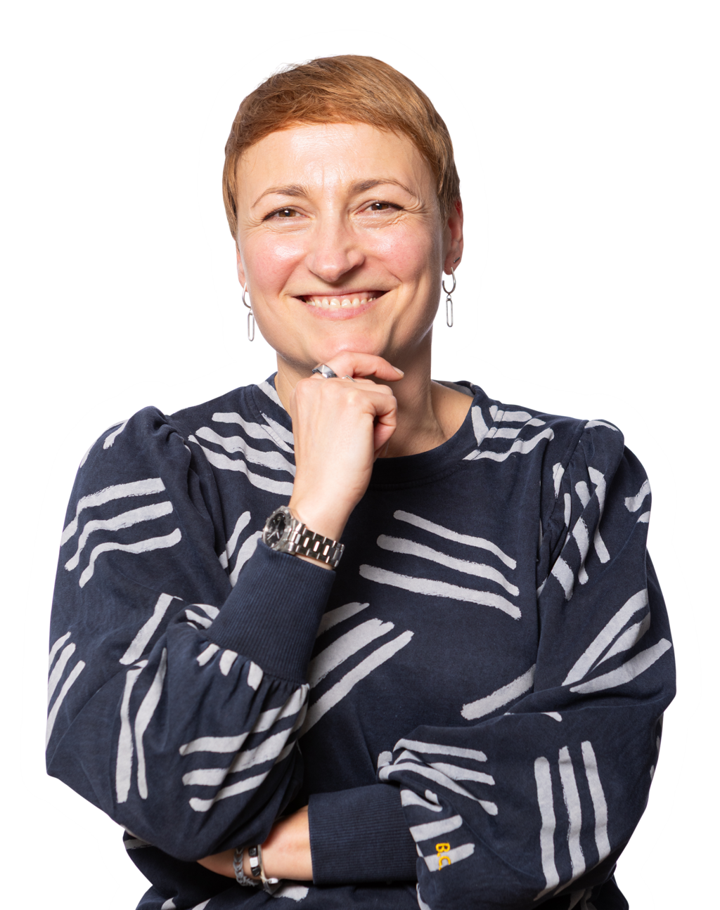 A smiling woman with short, reddish-brown hair and orange pants, wearing a dark blue shirt with white stripes, poses with her hand on her chin.