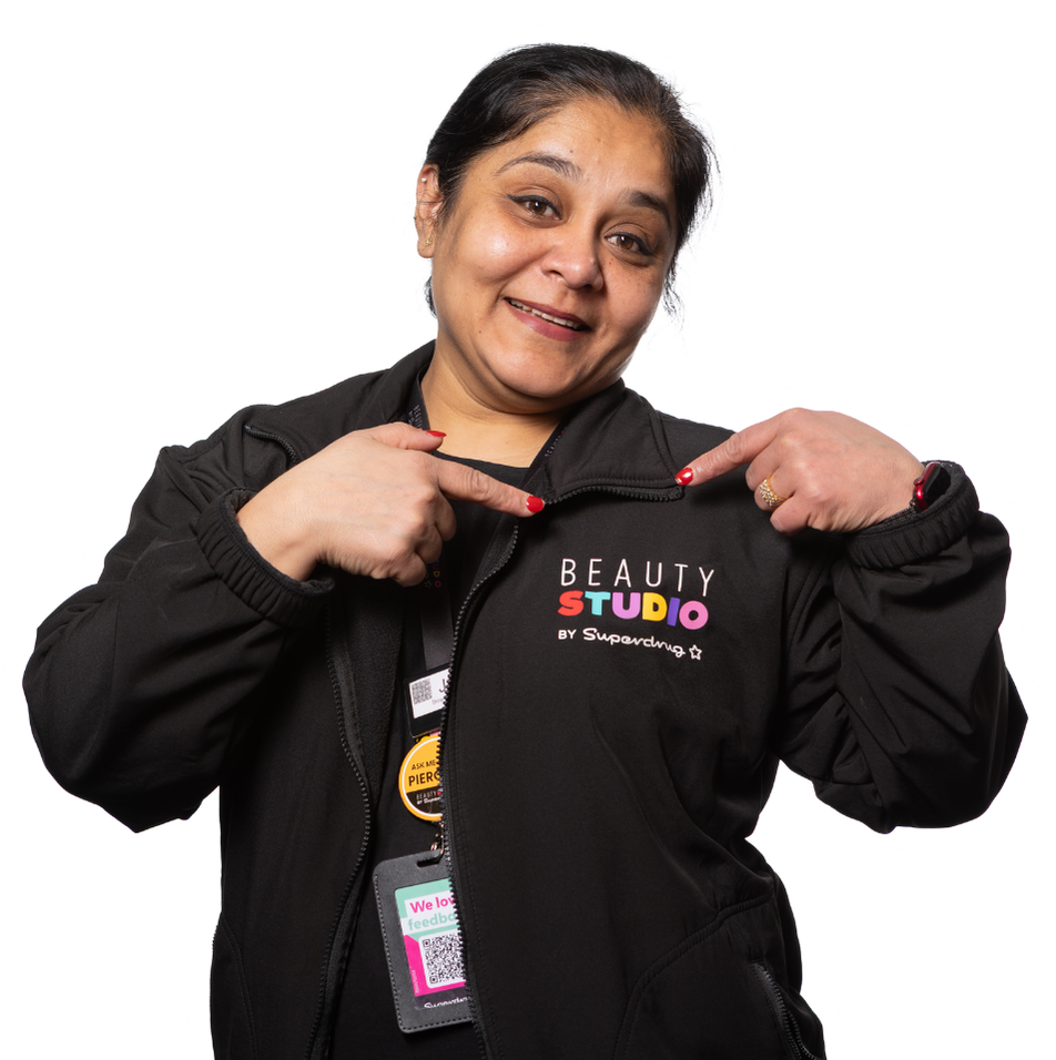 A smiling woman points to her "Beauty Studio by Superdrug" jacket, wearing a lanyard with multiple tags.