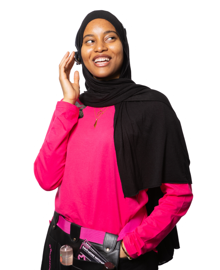 A Muslim woman wearing a hijab and a makeup apron, with makeup products, smiles and looks up while holding her ear.